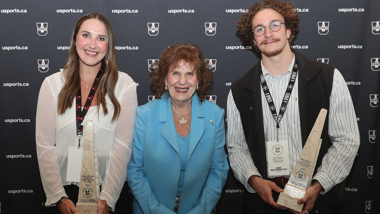 Three individuals posing in front of a U SPORTS branded backdrop, with Sarah Gates and Gabe Mastromatteo on the left and right sides, respectively, holding their athlete of the year trophies