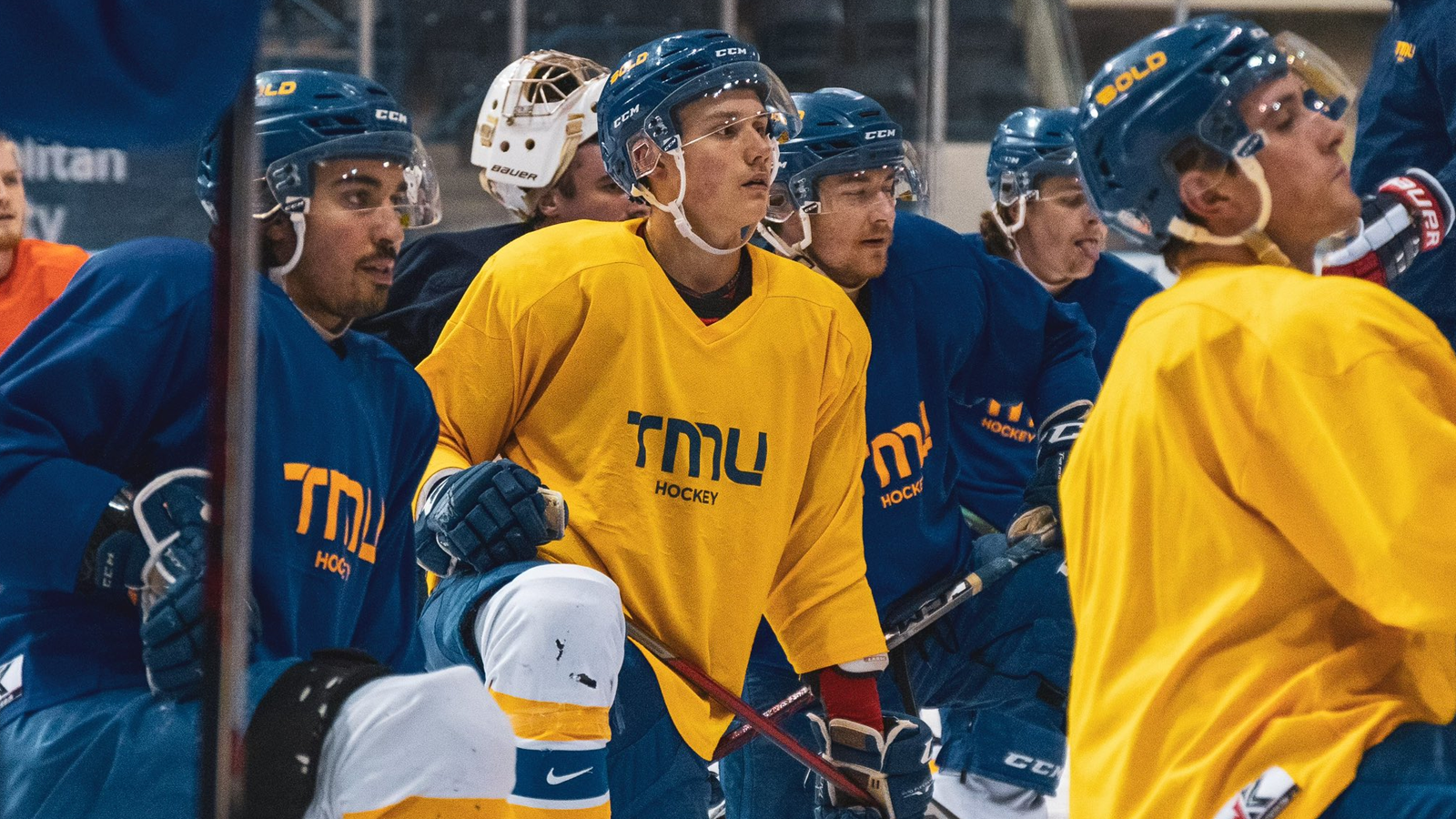 TMU men's hockey team on the ice, while taking a knee, as they look onward at their coach during a practice
