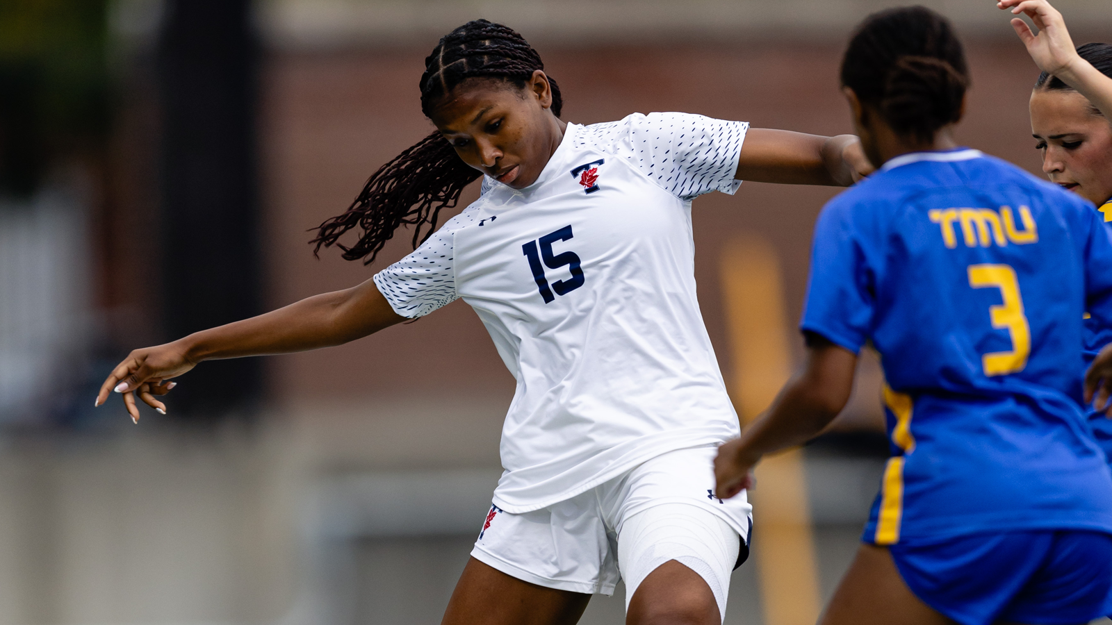 Toronto Varsity Blues women's soccer player looking downward while kicking the ball, with a TMU player defending in front of her